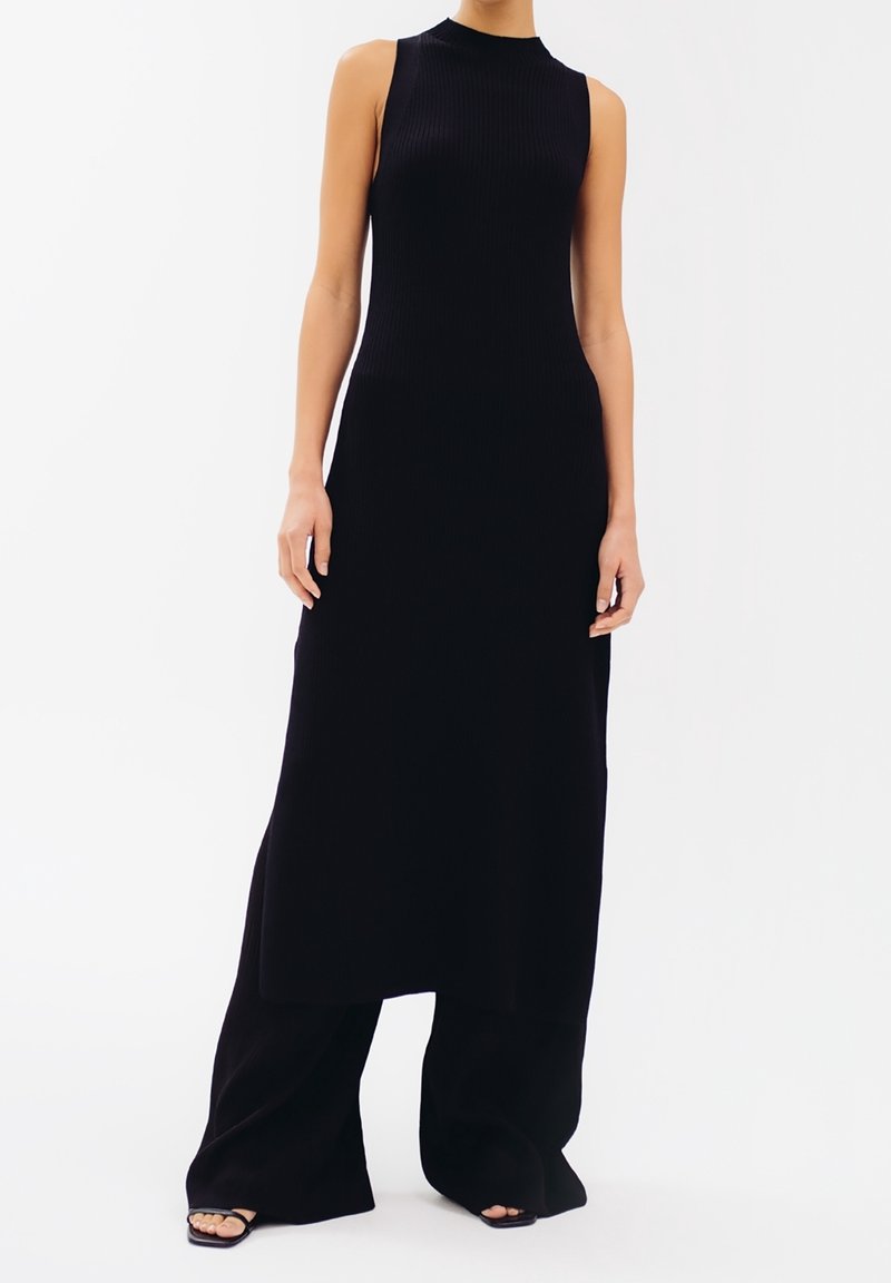 Abito lungo nero a coste con collo alto e design senza maniche. Nella parte inferiore presenta pantaloni larghi e fluenti. Sono indossati sandali neri flat.