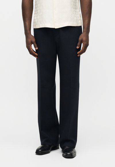 Άνδρας που φοράει μαύρο παντελόνι με ίσια γραμμή, μαύρα παπούτσια και εκρού κοντομάνικο πουκάμισο μέσα, στέκεται μπροστά σε απλό φόντο.