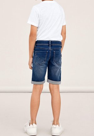 Kind draagt opgerolde donkerblauwe spijkershorts, wit T-shirt en witte sneakers, staat met de rug naar de camera gericht.