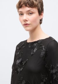 Μαύρο τοπ με υφή κεντητού λουλουδιού, στρογγυλή λαιμόκοψη και ελαφρύ ύφασμα. Το σχέδιο περιλαμβάνει περίπλοκες, ανάγλυφες λουλουδένιες λεπτομέρειες.