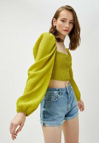 Gul tekstureret crop top med pufærmer, parret med slidte lyseblå denimshorts. Fremtrædende samlinger og elastiske detaljer er synlige.