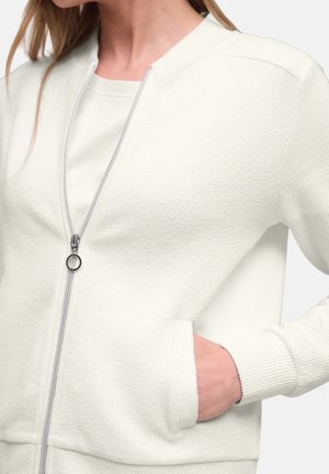 Donna che indossa una giacca zip-up testurizzata color avorio con cerniera a tirante ad anello e mano nella tasca laterale, vista dal collo ai fianchi.