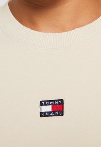 Primo piano di una maglietta beige a girocollo con una piccola toppa rettangolare del logo Tommy Jeans cucita sul petto.