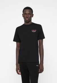 Fekete pamut póló, amelyen egy kis rózsaszín betűs logó található: "CLSR LDN" a mellrészen. Kerek nyakkivágású, rövid ujjak, testhezálló fazon.