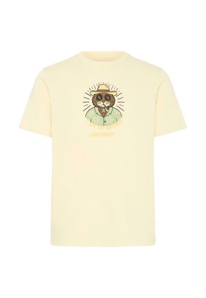 T-shirt couleur crème avec un graphisme représentant un lion portant des lunettes de soleil, un chapeau et une chemise fleurie, accompagné du texte « TENUE HORS BUREAU ».