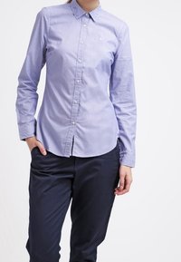 Person standing with left hand in pocket, wearing a light blue button-up shirt and dark navy pants against a plain background.