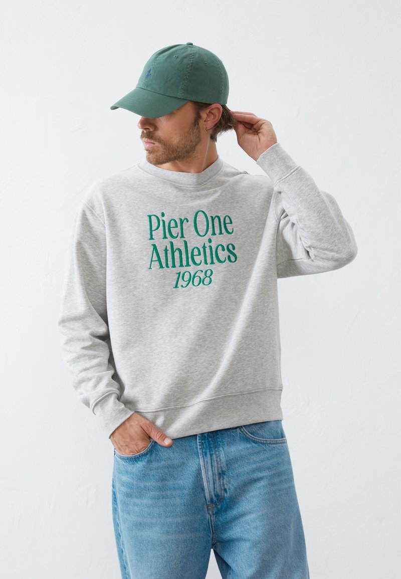 Szürke pulóver "Pier One Athletics 1968" felirattal zöld színben, világoskék farmernadrággal és zöld sapkával párosítva. Puha anyag, laza szabású.
