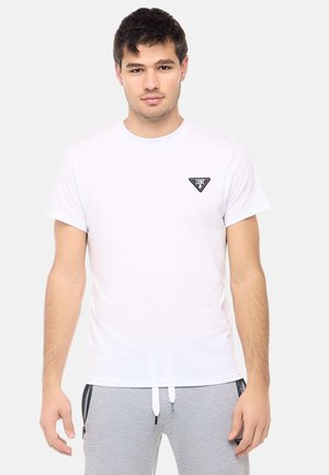 Jonge man die een effen wit T-shirt draagt met een klein driehoekig logo op de borst, gecombineerd met lichtgrijze joggingbroeken.