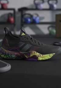 Chaussure de sport noire avec semelle multicolore à motifs, posée sur le sol de la salle de sport, avec des kettlebells de différentes couleurs floutées en arrière-plan.