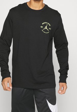 Homme portant un t-shirt noir à manches longues avec un "Mike's Gym" jaune et un logo de joueur de basket-ball sur la poitrine gauche, associé à un short de sport noir et blanc.