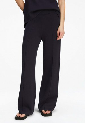 Frau trägt schwarze weit geschnittene Hosen und schwarze Sandalen mit quadratischen Zehen, steht vor einem weißen Hintergrund.