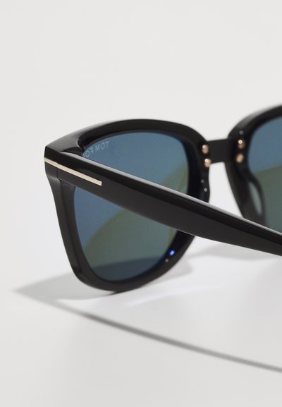 Black sunglasses with blue-tinted lenses. Bold, angular frame featuring a silver stripe on the side and small decorative studs on hinges.