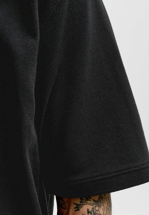 Svart teksturert kortermet stoff med en delvis synlig tatovert underarm under ermet mot en ensfarget lys bakgrunn.