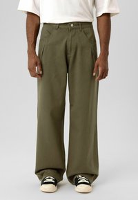 Olijfgroene wijde broek van stevig stof, met steekzakken aan de voorkant en een knoopsluiting, gecombineerd met zwart-witte sneakers.