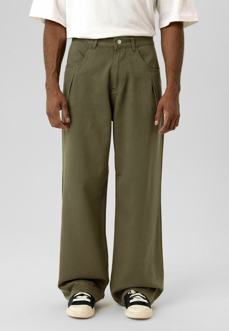 Olijfgroene wijde broek van stevig stof, met steekzakken aan de voorkant en een knoopsluiting, gecombineerd met zwart-witte sneakers.