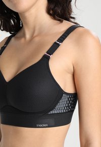 Svart sports-bh med mesh-struktur, justerbare stropper og rosa detaljer. Har et mønstrete sidelag og et merket bånd nederst.