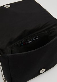 Sort stofpose med klaplukning og sølvdetaljer. Indersiden har en etiket, tekstureret overflade og ingen synlige mønstre.