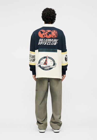 Άνδρας που φοράει μπλε σκούρο και κρεμ μακρυμάνικο πουκάμισο με γραφικά Billionaire Boys Club και θεματολογία ιστιοπλοΐας, συνδυασμένο με χαλαρά γκρι παντελόνια και αθλητικά παπούτσια.