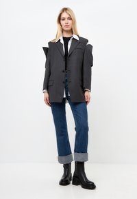 Grijze oversized blazer met hoekige schouderaccenten, zwarte knopen en een opgestikte zak, gecombineerd met blauwe oproljeans en zwarte enkellaarzen.