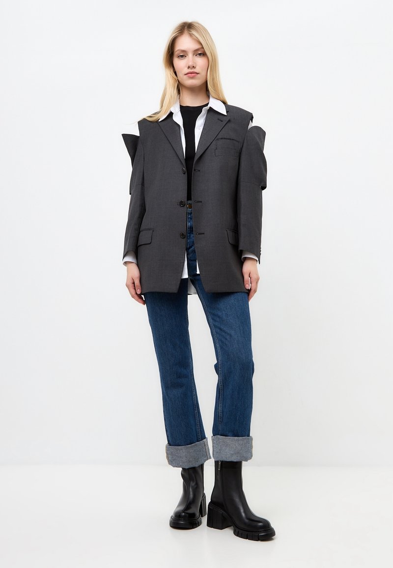 Grijze oversized blazer met hoekige schouderaccenten, zwarte knopen en een opgestikte zak, gecombineerd met blauwe oproljeans en zwarte enkellaarzen.
