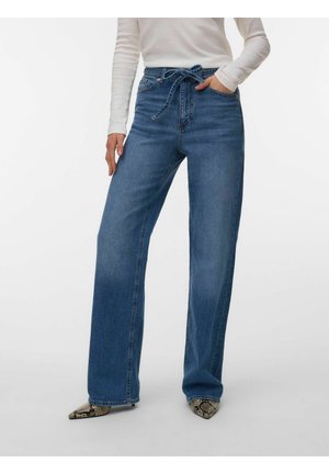 Kvinne iført blå vidbukser med høyt liv og knytt belte, hvit langermet topp og spisse sko med slangeskinnmønster.