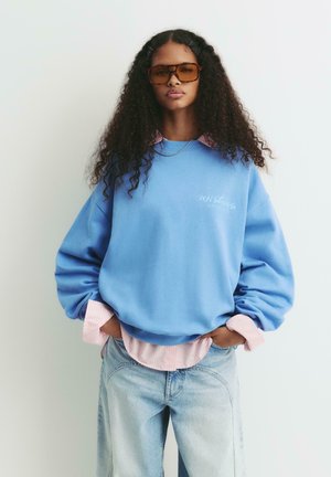 Felpa blu oversize con polsini a costine, indossata sopra una maglietta a righe rosa; abbinata a jeans azzurri a vestibilità rilassata e occhiali da sole grandi.