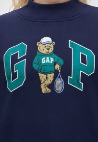 Ours portant une casquette et un sweat à capuche GAP, appuyé sur une raquette de tennis entre les grandes lettres G et P sur un tissu bleu marine.