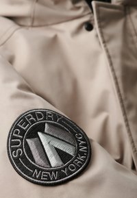 Beige vandtæt jakke med en tekstureret stof. Har et cirkulært sort broderet patch med teksten "Superdry" og "New York, NYC" samt et geometrisk design.