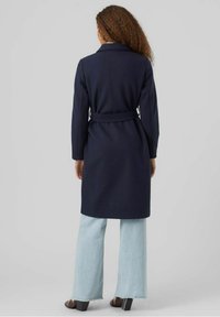 Cappotto blu navy con maniche lunghe e vita con cintura, abbinato a jeans a gamba larga di un azzurro chiaro. Il cappotto presenta un colletto a punta e un design strutturato.