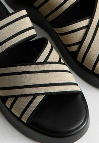 Zwarte en beige gestreepte stoffen sandalen met gekruiste banden, een gevoerde zwarte zool en een plat ontwerp voor comfort.