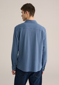 Camisa azul de manga larga con botones, confeccionada en tela texturizada, con cuello y puños de un solo botón, llevada sobre pantalones vaqueros oscuros.