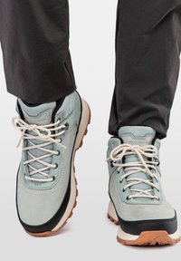 Zapatos de senderismo azul claro con materiales de ante y malla, cordones beige, suelas de goma y acentos negros. Diseñados para actividades al aire libre.