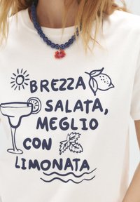 T-shirt blanc avec un texte graphique bleu et des illustrations d'un soleil, d'un cocktail, d'un citron et de feuilles de menthe. Collier de perles bleu rond avec un pendentif en forme de crabe.