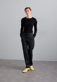 Black long-sleeve top, tailored black trousers, and yellow leather shoes with a low heel, standing on a light gray floor.