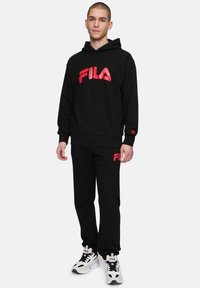 Sudadera negra con capucha y pantalones de chándal con un gran logo rojo de FILA. El material es una mezcla de algodón. Las zapatillas tienen un diseño blanco y negro con cierre de cordones.