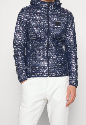 Marineblaue gesteppte Jacke mit Kapuze, silberne EA7-Logos überall, durchgehender Frontreißverschluss, getragen mit weißen Hosen, Hände in den Taschen.