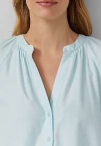 Camicetta azzurro chiaro a maniche corte con scollo rotondo e leggere arricciature, dotata di una placket con bottoni lungo la parte anteriore. Texture liscia.