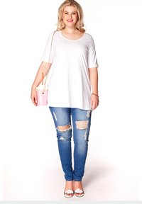 Witte oversized t-shirt, blauwe ripped skinny jeans en witte sandalen met tekst. Houdt een roze beker vast met een rietje en kettingstrap.