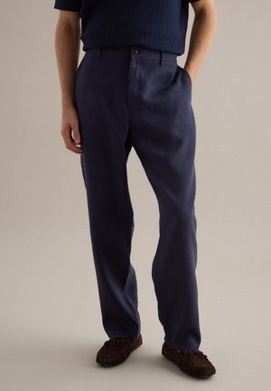 Man draagt donkerblauwe broek, marineblauw geribd T-shirt met korte mouwen en bruine suède mocassins, staat met één hand in de zak.