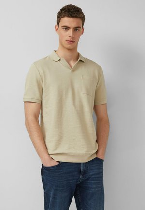 Junger Mann mit kurzen lockigen Haaren, der ein beiges, kurzärmeliges, strukturiertes Polohemd und blaue Jeans trägt, die Hände in den Taschen, vor einem hellgrauen Hintergrund.