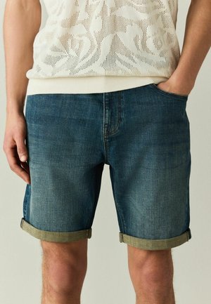 Mann trägt hochgekrempelte dunkelblaue Jeansshorts und ein weiß gemustertes Strickoberteil, eine Hand in der Tasche, vor einfarbigem Hintergrund.