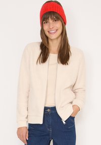 Beżowa, pluszowa kurtka zapinana na zamek z prążkowanymi mankietami i kołnierzem. Modelka ma na sobie jasną bluzkę i czerwony, dziergany opaska na głowę. Ciemnoniebieskie dżinsy.