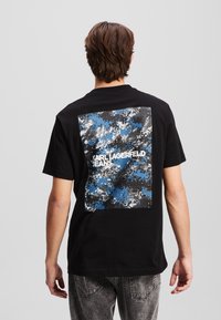 Juodas medvilninis marškinėlis su mėlynu ir pilku pikseliuotu grafiku nugaros pusėje, kuriame baltais rašmenimis užrašyta "KARL LAGERFELD JEANS". Standartinio kirpimo.