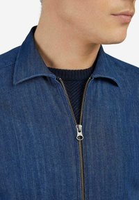 Giacca di jeans con colletto strutturato, dotata di una zip metallica a tutta lunghezza e di un maglione a trama blu scuro sotto.