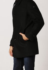 Cappotto di lana nera con design doppiopetto, grandi bottoni, colletto a revers e tasche laterali. Texture liscia con vestibilità su misura.