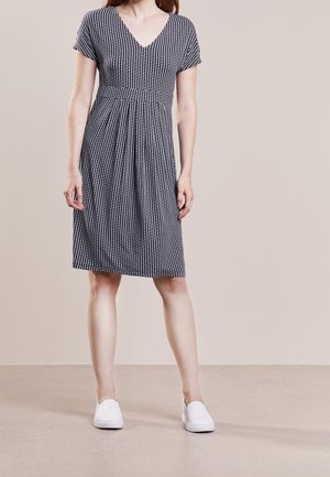 Woman standing wearing a knee-length short-sleeve black and white patterned dress with white slip-on shoes on a plain floor.