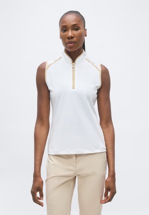 Frau steht frontal, trägt ein weißes ärmelloses Zip-up-Top mit beigem Besatz und beige Hosen vor einem schlichten weißen Hintergrund.