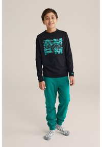 Čierna dlhá košeľa s tyrkysovým textom, spárovaná so zelenými teplákmi. Model má na sebe šedé a biele kostkované ponožky.