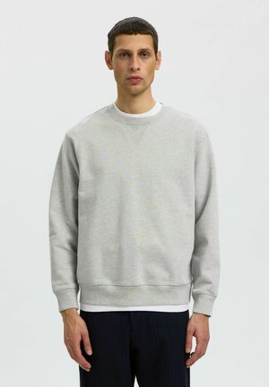 Homme portant un sweat-shirt gris clair à col rond par-dessus une chemise blanche et un pantalon foncé, debout devant un fond clair uni.