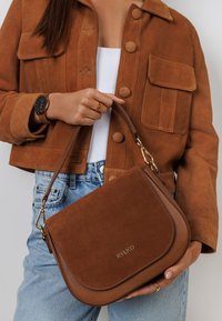 Bruine suede handtas met een gebogen vorm en gouden hardware. Subtiele logo aan de voorkant, vastgehouden door een hand tegen een bruine jas en denim.
